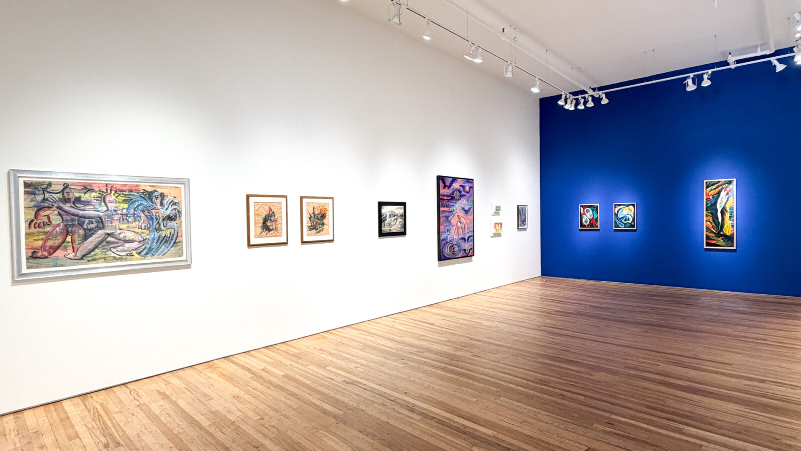 白い壁に大小の絵画が並ぶ展示室。突き当たりの壁は濃い青で、色彩の強い抽象画が3点掛かっている
