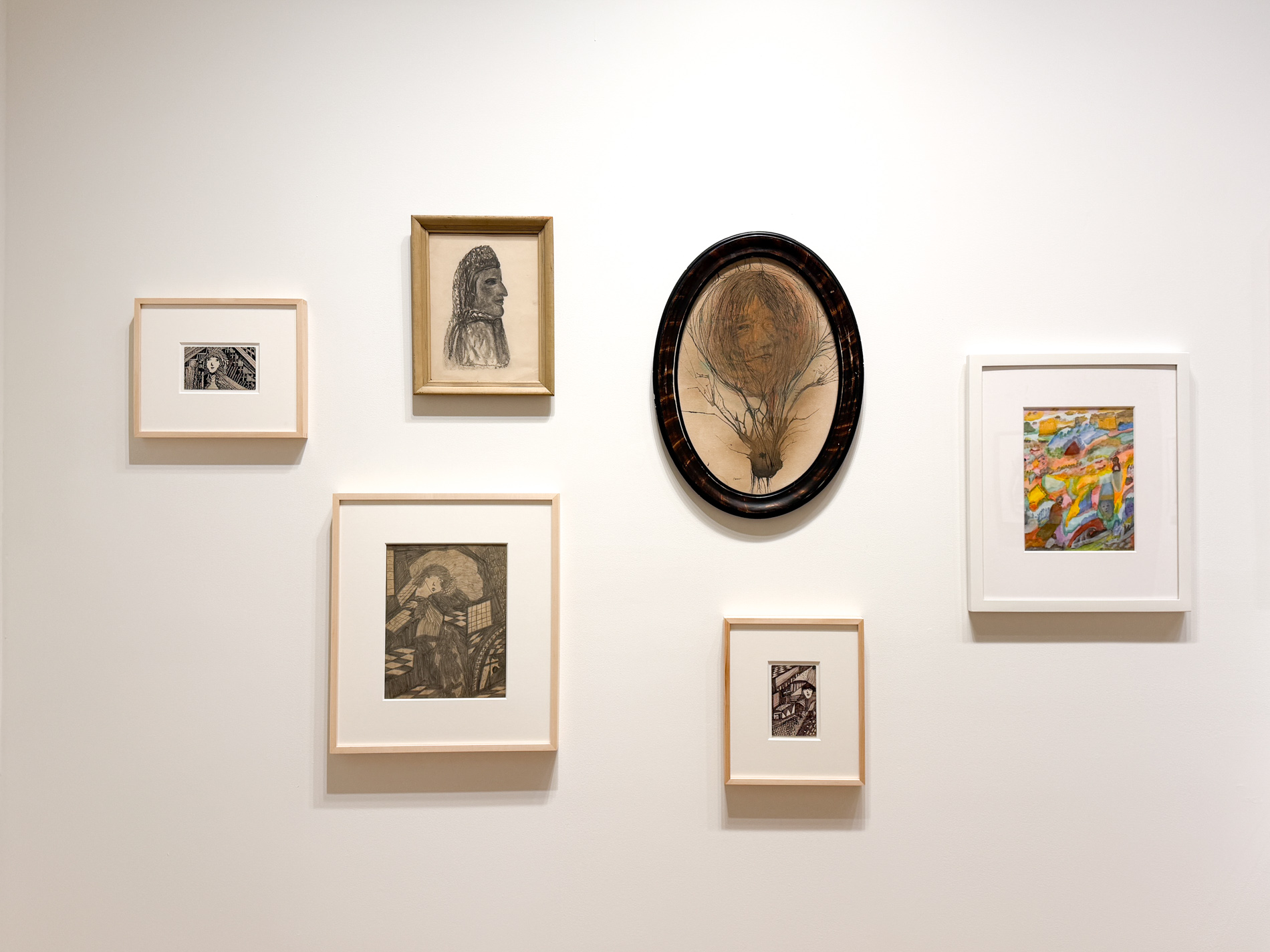 白い壁に小さな額が6点。白黒のインク画、横顔の素描、楕円の額に入った絵、色鮮やかな小品など