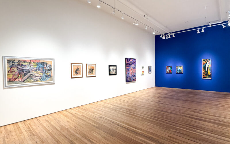 白い壁に大小の絵画が並ぶ展示室。突き当たりの壁は濃い青で、色彩の強い抽象画が3点掛かっている