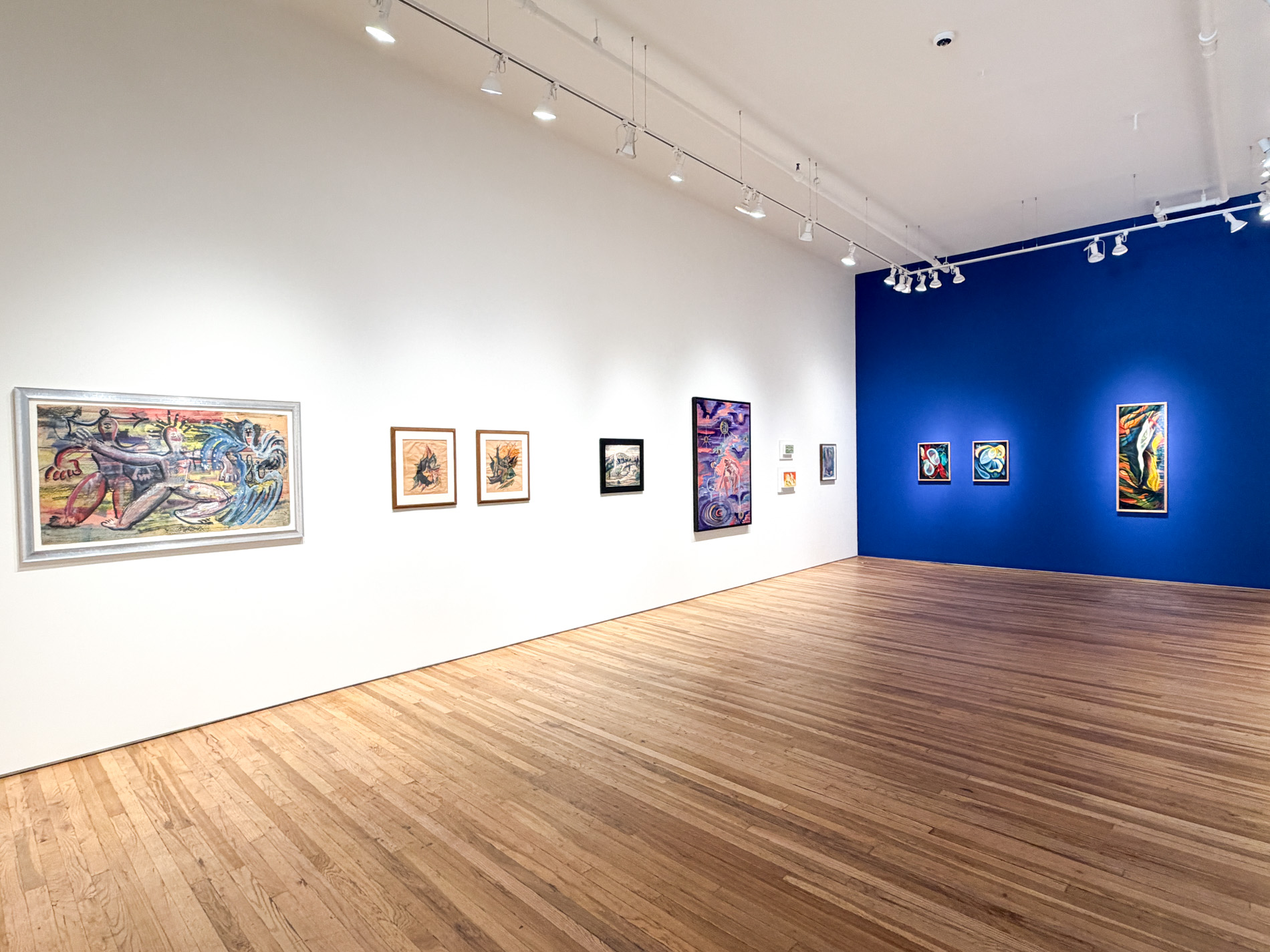 白い壁に大小の絵画が並ぶ展示室。突き当たりの壁は濃い青で、色彩の強い抽象画が3点掛かっている