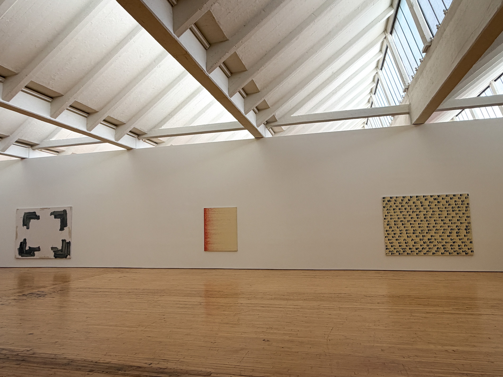 天窓の下の白い壁に絵画が3点、間隔を空けて掛かっている。左は四隅に灰色の筆跡、中央は淡い色の縦長の画面、右は黄土色の地に筆の点が規則正しく並ぶ