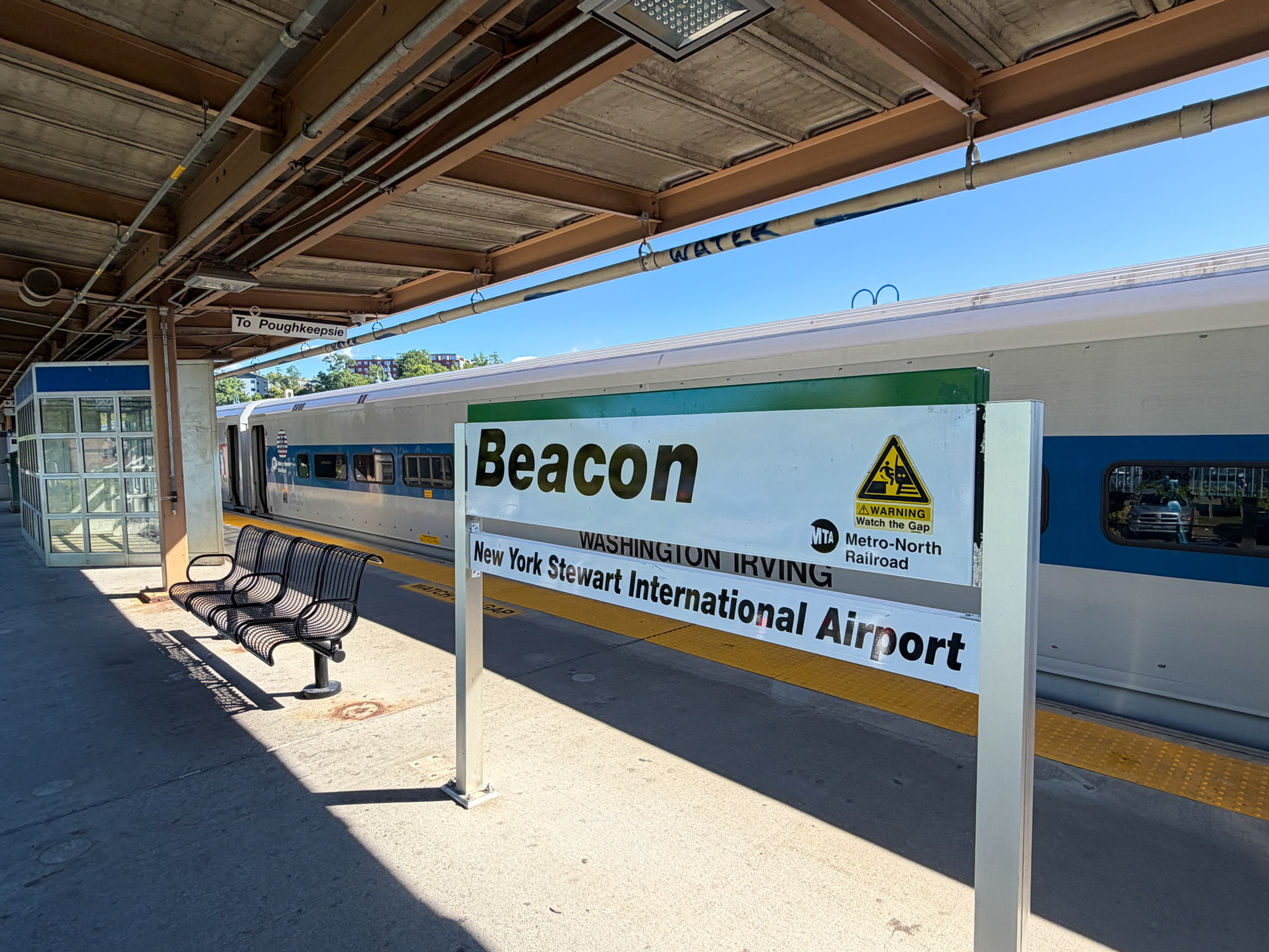 駅のホームに立つ「Beacon」の駅名標。屋根の下にベンチが並び、奥に銀色の車両が停まっている
