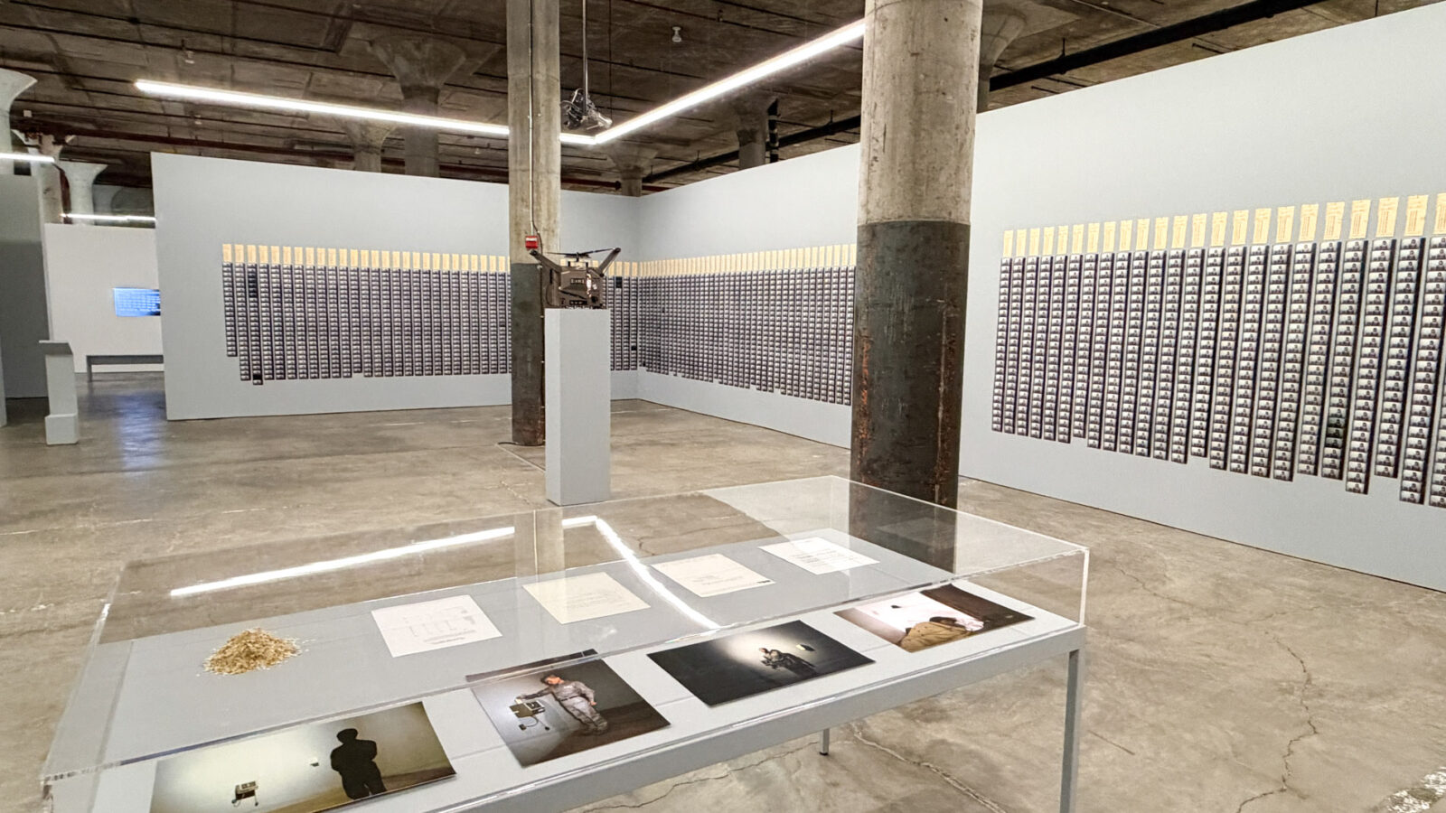 コンクリートの柱が並ぶ地下の展示室。壁一面にタイムカードと写真のコマが並び、手前にガラスケースが置かれている
