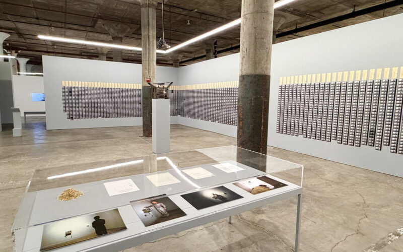 コンクリートの柱が並ぶ地下の展示室。壁一面にタイムカードと写真のコマが並び、手前にガラスケースが置かれている