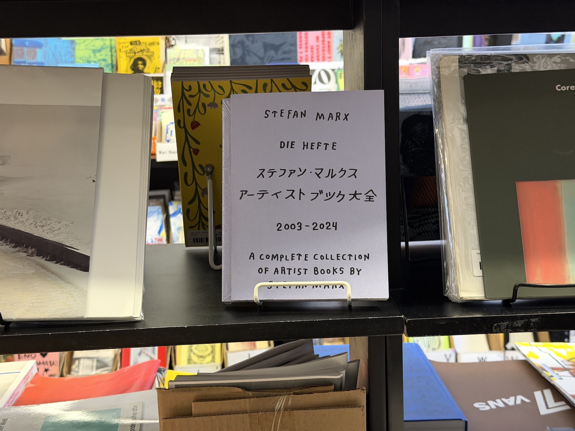 白い表紙に手書き風の文字で「STEFAN MARX DIE HEFTE ステファン・マルクス アーティストブック大全 2003–2024」と書かれた本が、書見台に立てられている