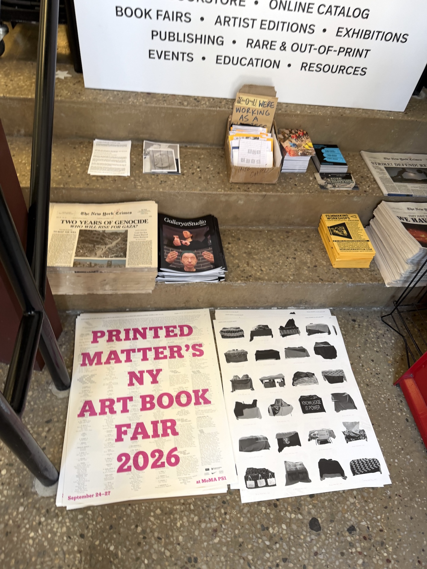 コンクリートの階段の下に、ピンクの文字で「PRINTED MATTER'S NY ART BOOK FAIR 2026」と書かれたポスターが置かれ、段の上に新聞や冊子が並ぶ