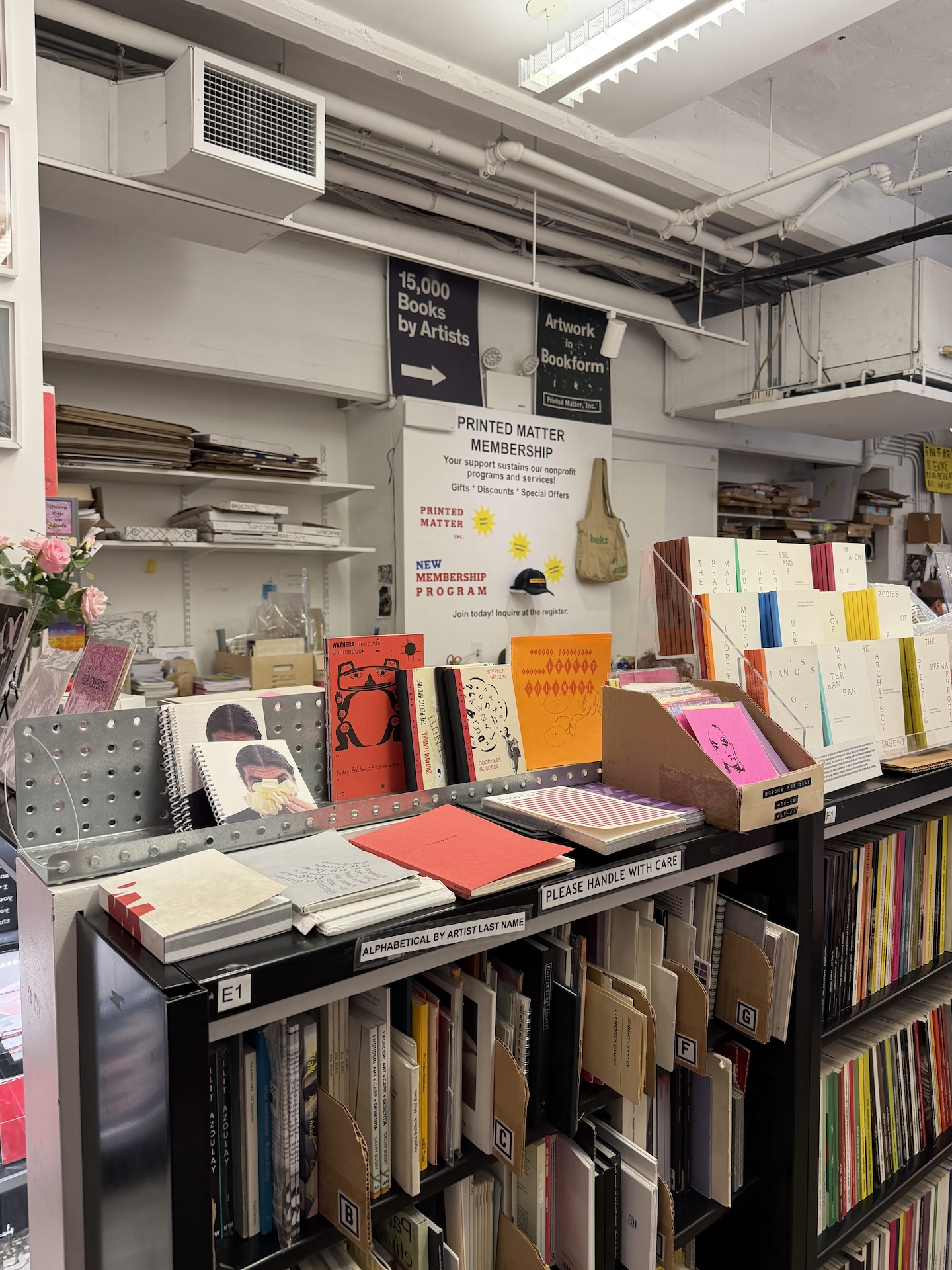 天井近くに「15,000 Books by Artists」「Artwork in Bookform」と書かれた黒い案内板。その下に会員募集の看板と、色とりどりの冊子を並べた棚