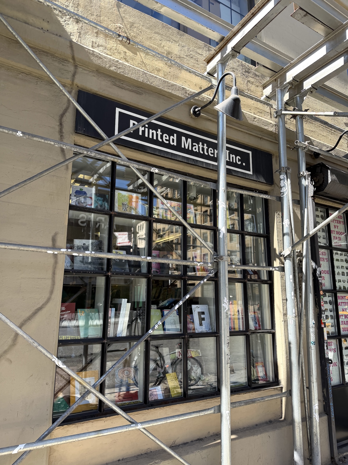 黒地に白文字で「Printed Matter, Inc.」と書かれた看板の下に、格子状の大きな窓。窓の内側に本や冊子が飾られ、手前を足場のパイプが横切っている