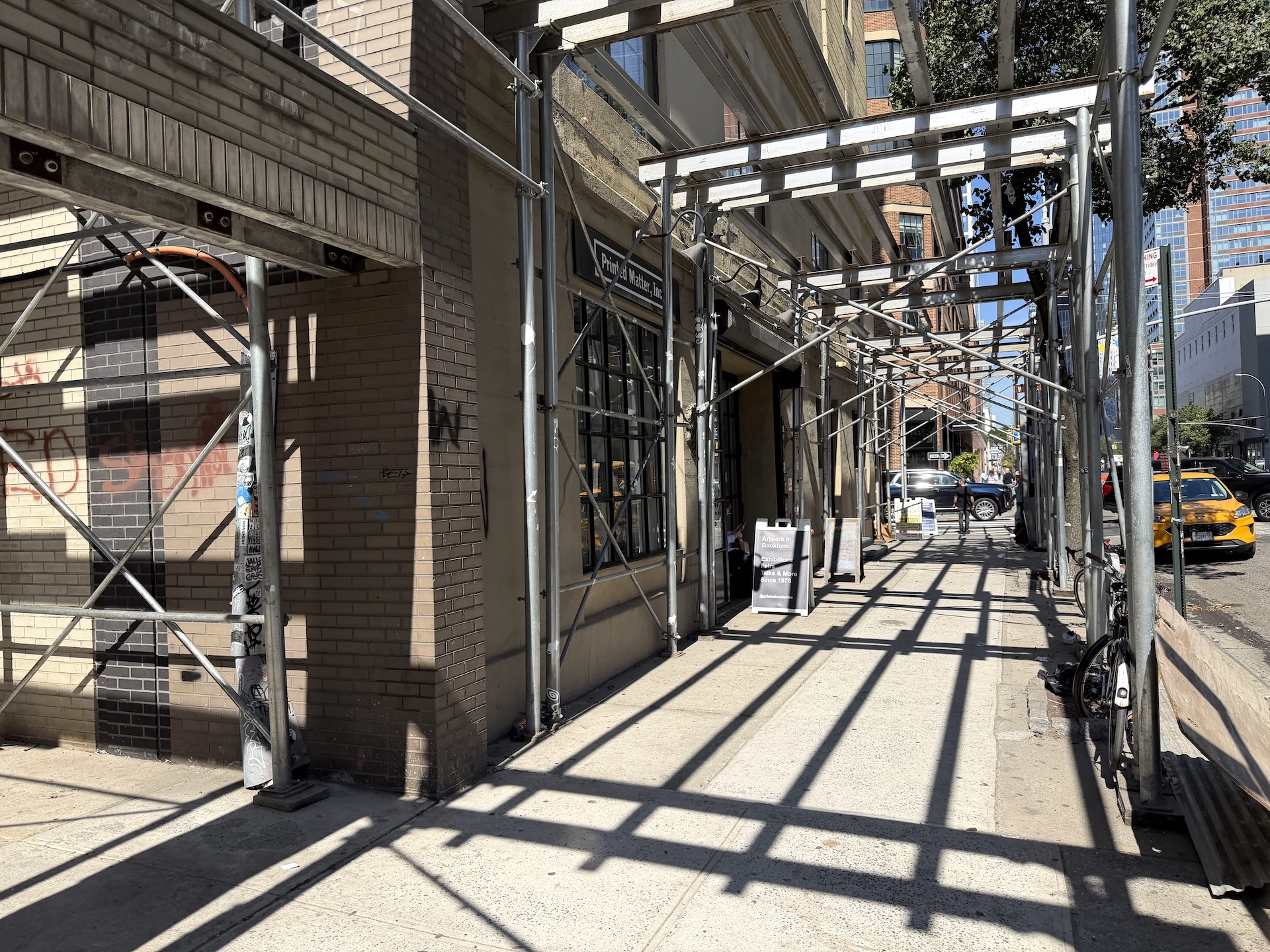 足場に覆われた歩道。建物の1階に黒い看板と格子窓があり、店先に立て看板が2つ置かれている。奥の車道には黄色いタクシー