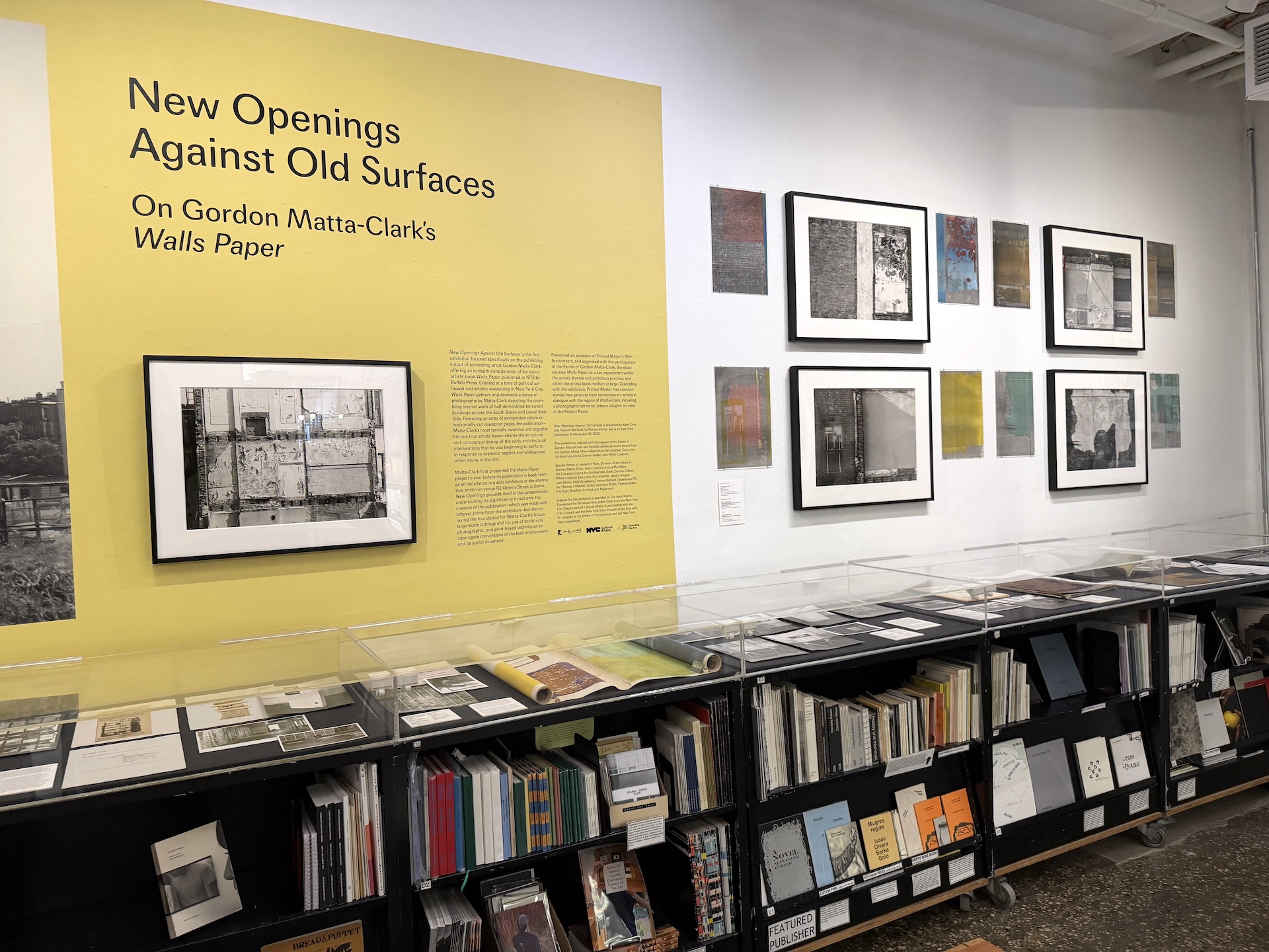 黄色い壁に展覧会タイトルと解説文、額装された白黒写真。右の白い壁に色のついたプリントと写真が並び、手前にガラスケースと本棚