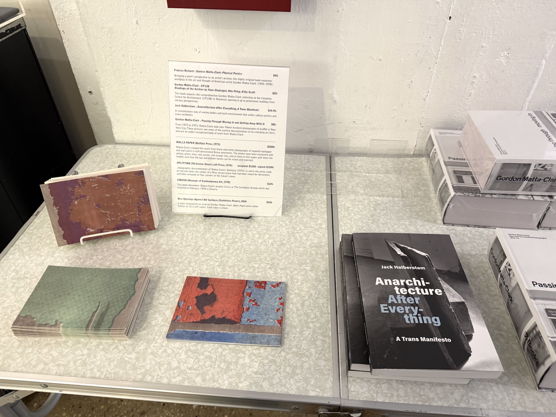 白い台の上に、剥がれた壁を写したカラーの冊子や、黒い表紙の本『Anarchitecture After Everything』が置かれ、奥に英文の価格表が立っている