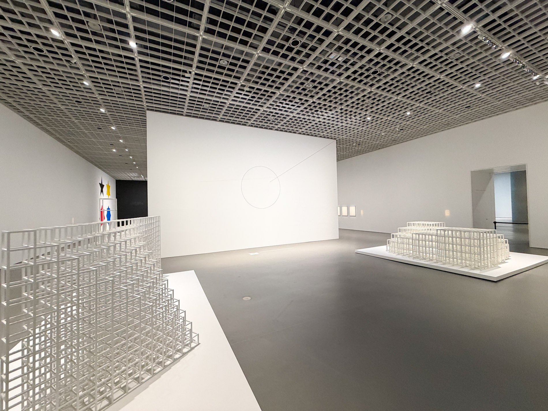 白い格子状のモジュール構造物が展示室の床の台の上に大きく展開し、奥にも別の白い構造物が置かれている