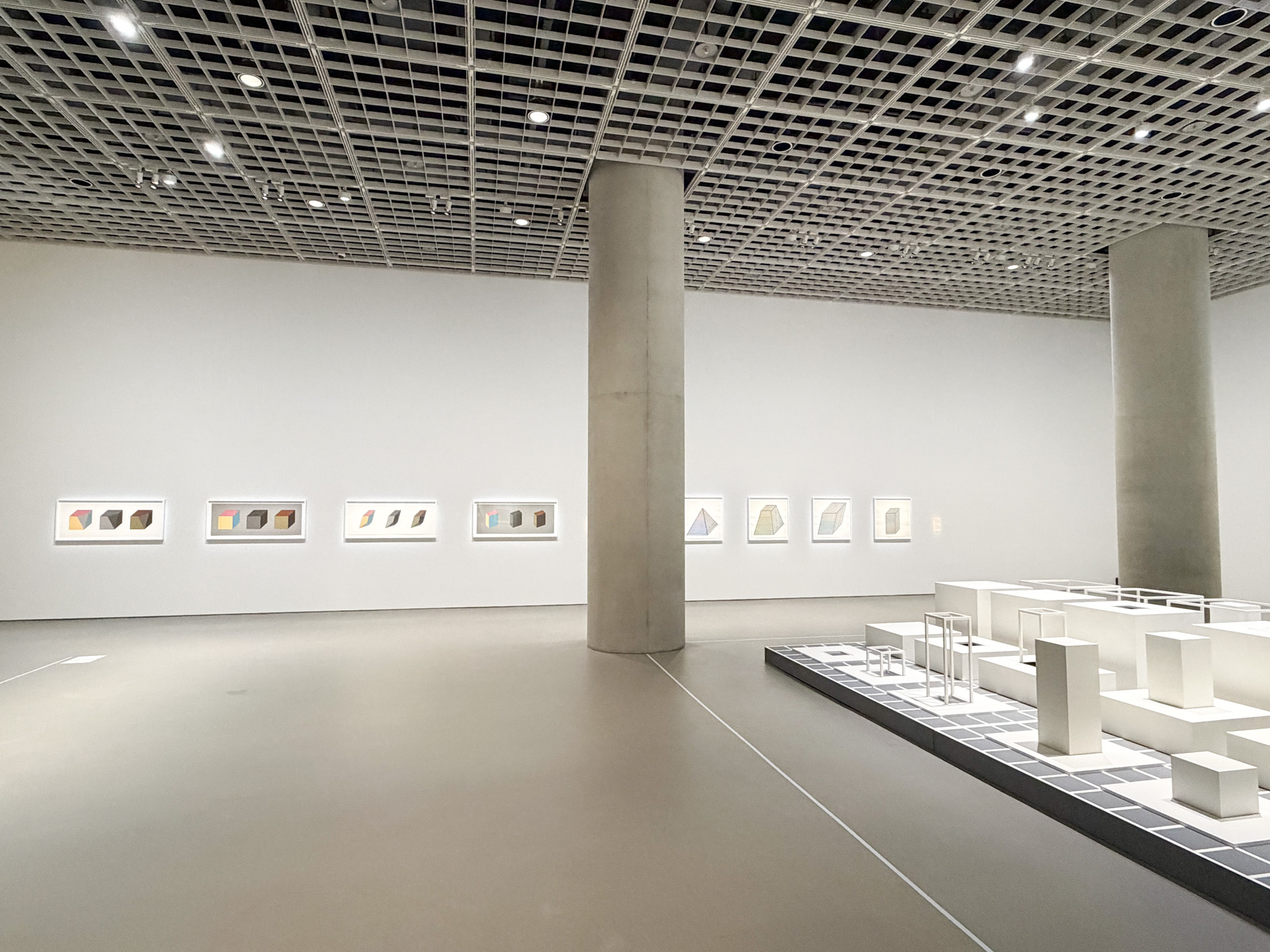 展示室の壁一面に小さな額装のドローイングと版画が等間隔で並び、右手前の低い台に白い構造物の群が置かれている