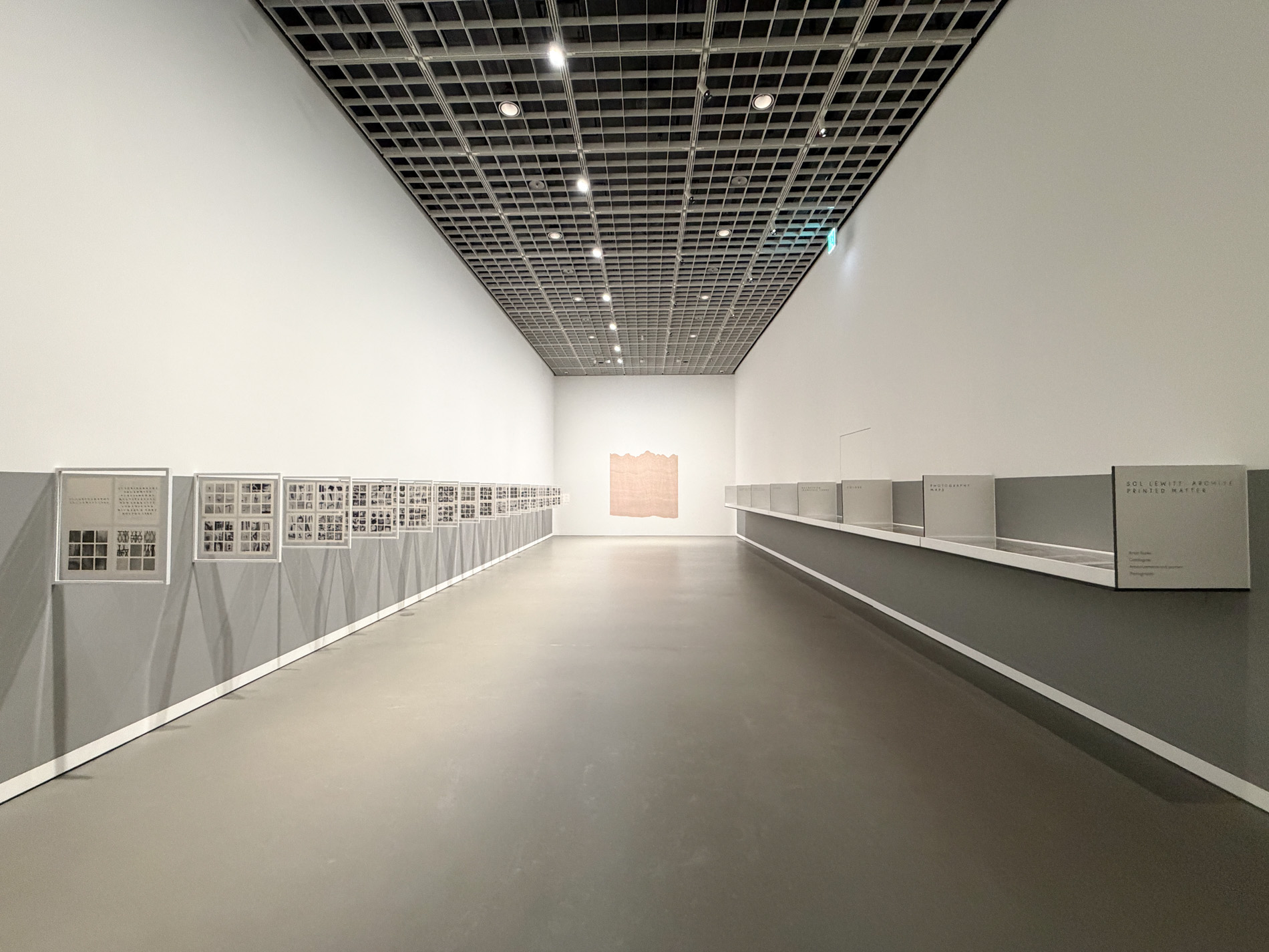 細長い回廊。左の壁には本の見開きを額装した小さな作品が斜めに突き出して長く連なり、右手には低い展示棚が奥まで続く。突き当たりの壁に淡いピンクの作品が一点掛かる