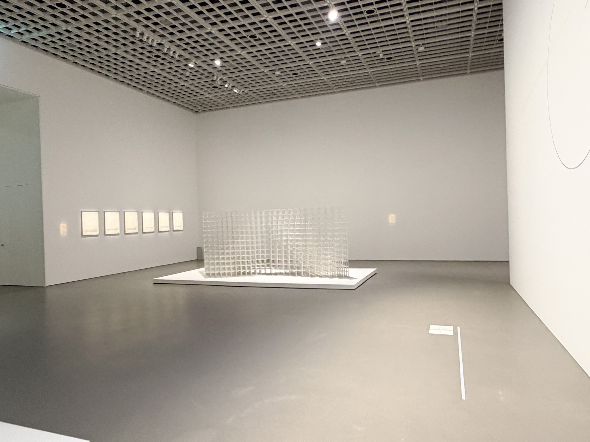 白い展示室の中央に、開いた立方体を格子状に組み上げた白い構造物が低い台の上に据えられ、左手の壁には小さな額装作品が一列に並ぶ