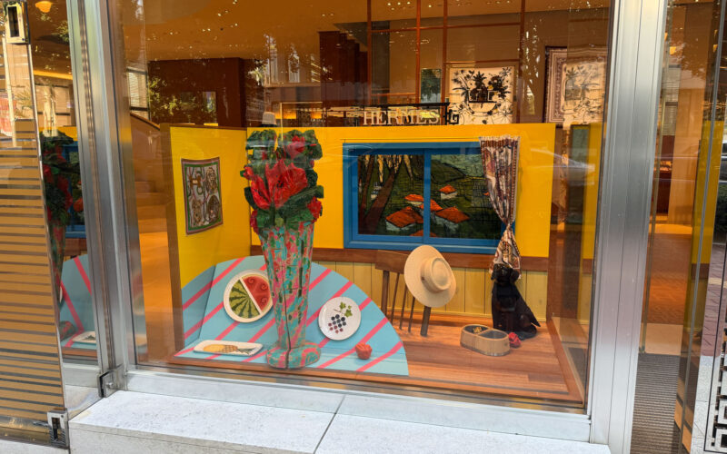 黄色い壁の室内を模したウィンドウ。木彫の花瓶に赤い花、水色のテーブルにスイカの皿、青枠の窓の向こうに風景画