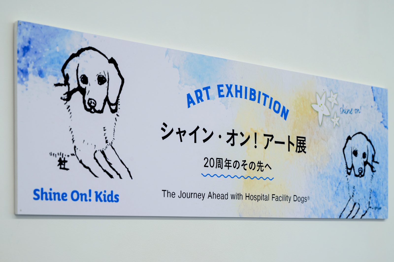 「シャイン・オン！アート展 20周年のその先へ」の展覧会サイン。水彩の青い背景に、線で描かれた2頭のホスピタル・ファシリティドッグ