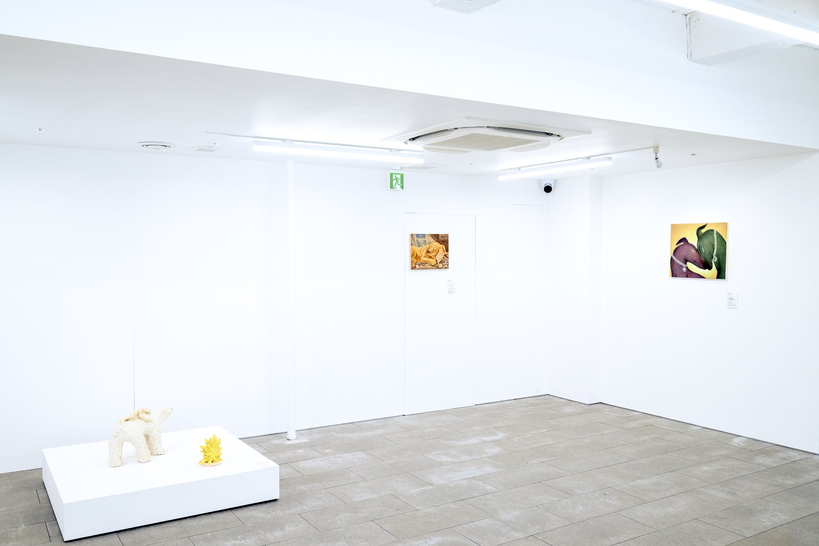 低い白い台の上に置かれた陶の犬と黄色い立体、奥の壁に絵画2点